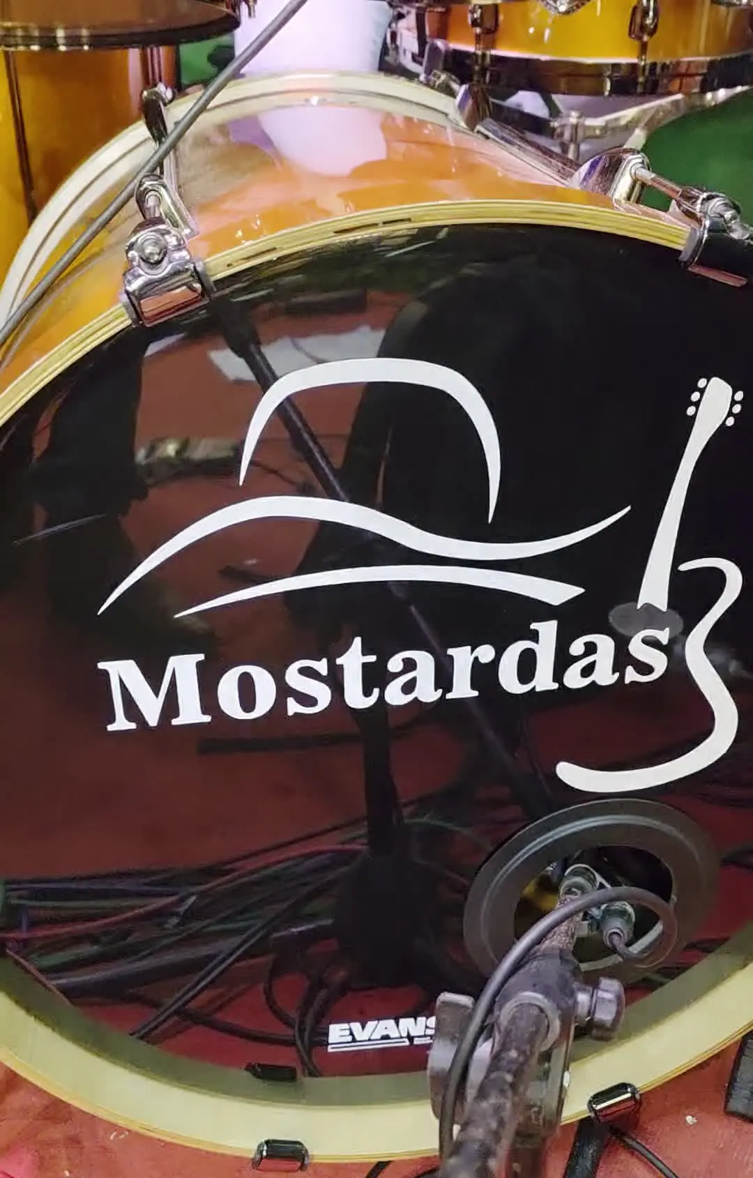 Bateria com o logótipo Mostardas no bombo, sobre carpete vermelha.
