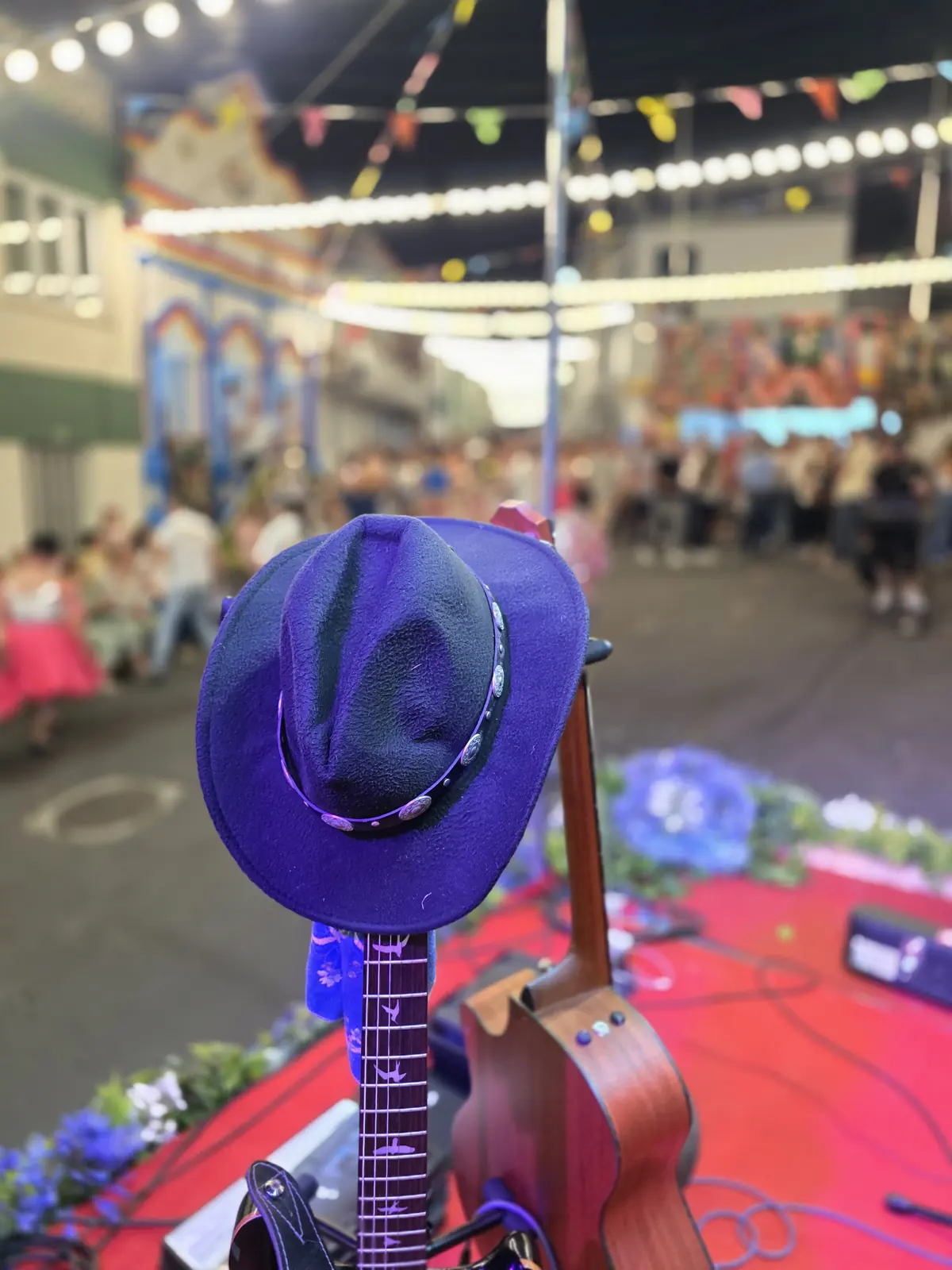 Chapéu de cowboy no palco e braço de guitarra, com o público da festa desfocado ao fundo.